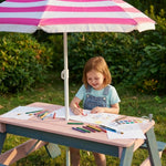 Child drawing at a table under a striped umbrella in an outdoor setting
